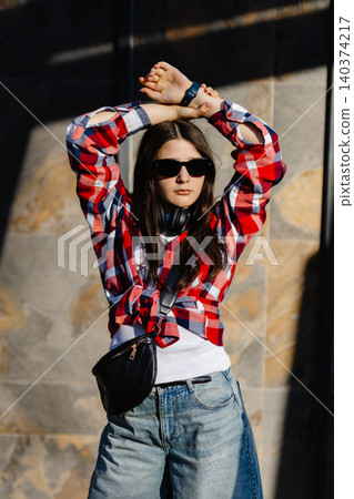 Stylish teen girl wearing sunglasses and headphones, posing against a wall 140374217