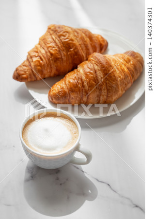Freshly brewed cappuccino coffee in a cup on a white marble background with croissants Freshly brewed cappuccino coffee in a cup on a white marble background with croissants 140374301