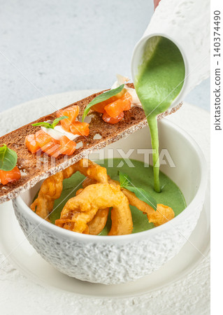 Pouring green herb cream soup with salmon crostini and crispy calamari in gourmet presentation 140374490