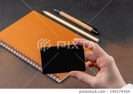 Hand holding blank black business card mockup over notebook on dark desk background branding identity template 140374504