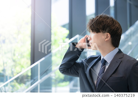 A young, handsome businessman is thinking while talking to a client on his smartphone by the window of an office building. A young, handsome businessman is thinking while talking to a client on his smartphone by the window of an office building. 140375219