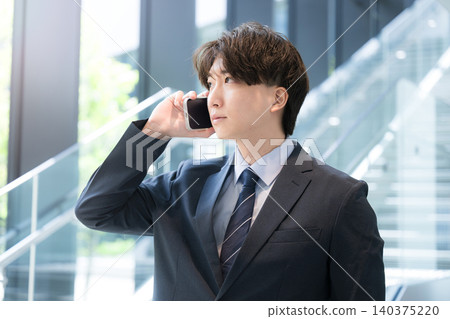 A young, handsome businessman is thinking while talking to a client on his smartphone by the window of an office building. A young, handsome businessman is thinking while talking to a client on his smartphone by the window of an office building. 140375220