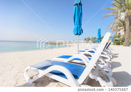 Beachside loungers arranged on a sunny day near calm waters and palm trees in a coastal paradise Beachside loungers arranged on a sunny day near calm waters and palm trees in a coastal paradise 140375887