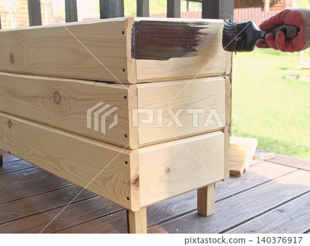 Brown oil for protection of wood by staining. Painting fence for protecting and pests. Woodworker applying finish to pine board box. Repairman varnishing with brush wooden fencing wall plank surface. Brown oil for protection of wood by staining. Painting fence for protecting and pests. Woodworker applying finish to pine board box. Repairman varnishing with brush wooden fencing wall plank surface. 140376917