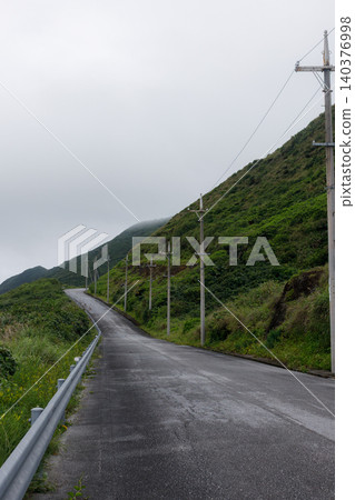 Under a cloudy sky, a wet road and utility poles stretch along the green mountainside. Under a cloudy sky, a wet road and utility poles stretch along the green mountainside. 140376998