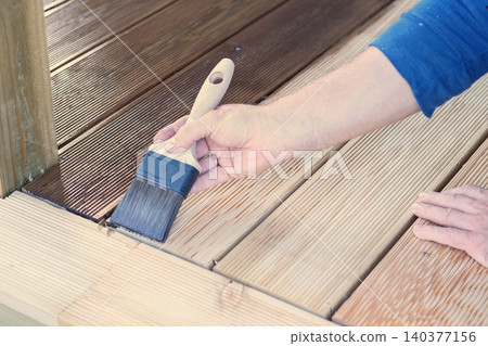 Brown oil for protection of wood by staining. Painting a larch for protecting and pests. Woodworker applying finish to terrace deck board. Repairman varnishing with brush wooden plank floor surface. 140377156