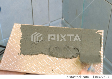 Tile adhesive spreading. Tiling mortar application. Worker smoothing adhesive. Workman smoothing grout on ceramic tiles. Bathroom renovation. Man using notched trowel, cement spread across tile back Tile adhesive spreading. Tiling mortar application. Worker smoothing adhesive. Workman smoothing grout on ceramic tiles. Bathroom renovation. Man using notched trowel, cement spread across tile back 140377307