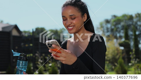 Young sport woman resting and using smartphone, drink water after workout in backyard. Sports female doing outdoor training. Young sport woman resting and using smartphone, drink water after workout in backyard. Sports female doing outdoor training. 140377696