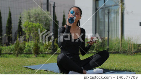 Young sport woman resting and using smartphone, drink water after workout in backyard. Sports female doing outdoor training. 140377709