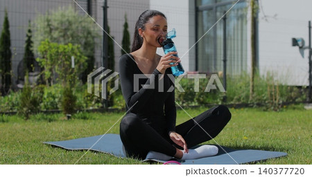 Young sport woman resting, drink water after workout in backyard. Sports female doing outdoor training. Young sport woman resting, drink water after workout in backyard. Sports female doing outdoor training. 140377720