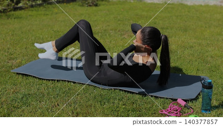Young woman makes press workout on the mat outdoor in sunny day. Woman in black sportswear doing exercise raising legs and pumping the press. Fitness training for burning fat calories. 140377857