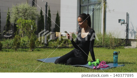 Young sport woman resting and using smartphone, after workout in backyard. Mobile application for training. Sports female doing outdoor training. Young sport woman resting and using smartphone, after workout in backyard. Mobile application for training. Sports female doing outdoor training. 140377895