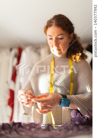 Craftswoman designer tailor adjusting fabric with measuring tape around her neck, focused on sewing details in a well-lit workshop with garments in the background Craftswoman designer tailor adjusting fabric with measuring tape around her neck, focused on sewing details in a well-lit workshop with garments in the background 140377992