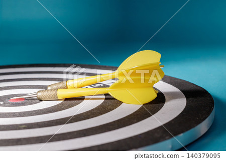 A close-up side angle shot of two yellow darts resting on top of a black and white circular dartboard, representing strategic pauses, planning, preparation, and project objectives. A close-up side angle shot of two yellow darts resting on top of a black and white circular dartboard, representing strategic pauses, planning, preparation, and project objectives. 140379095