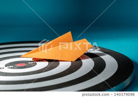 A low-angle close-up shot of an orange folded paper airplane resting on a circular dartboard, directed toward the target center, symbolizing a creative approach, direct marketing, and innovative A low-angle close-up shot of an orange folded paper airplane resting on a circular dartboard, directed toward the target center, symbolizing a creative approach, direct marketing, and innovative 140379096