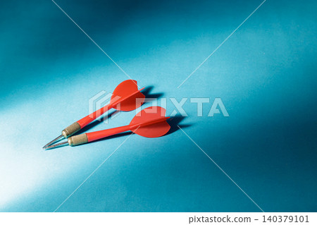 A pair of sharp red professional darts with metal tips lying parallel on a textured blue surface under dramatic side lighting, symbolizing targeting, focus, precision, and business strategy. 140379101