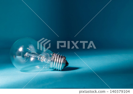 A clear filament lightbulb resting horizontally on a cyan surface with strong light and shadow contrast, serving as a classic symbol for business ideas, creativity, and technological innovation. A clear filament lightbulb resting horizontally on a cyan surface with strong light and shadow contrast, serving as a classic symbol for business ideas, creativity, and technological innovation. 140379104