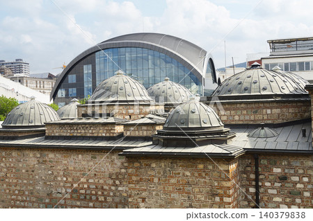 A historic stone building with domes is contrasted by a modern glass and steel structure in the background A historic stone building with domes is contrasted by a modern glass and steel structure in the background 140379838