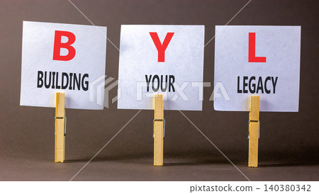 BYL building your legacy symbol. Concept words BYL building your legacy on beautiful white papers on clothespins. Beautiful grey background. Business BYL building your legacy concept. Copy space. BYL building your legacy symbol. Concept words BYL building your legacy on beautiful white papers on clothespins. Beautiful grey background. Business BYL building your legacy concept. Copy space. 140380342