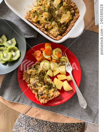 A health-focused meal features boiled potatoes, baked vegetables, fish, and broccoli pie served on a red plate. Kiwi slices are placed in a bowl. The setting is casual and inviting 140380828
