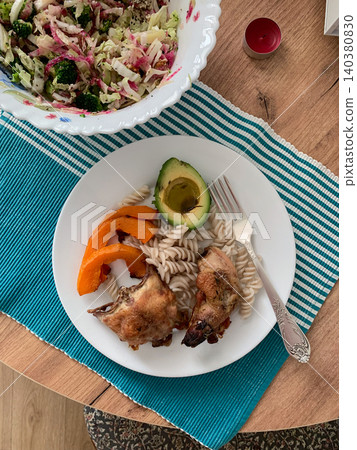 A bowl of fresh salad sits beside a plate of gluten-free pasta with sliced avocado, baked sweet potato, and chicken. The meal is arranged on a wooden table A bowl of fresh salad sits beside a plate of gluten-free pasta with sliced avocado, baked sweet potato, and chicken. The meal is arranged on a wooden table 140380830