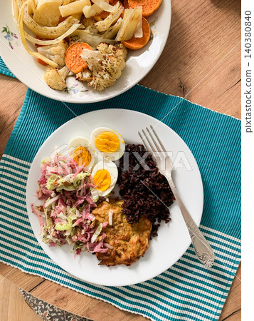 A plate features brown rice, boiled eggs, zucchini pancakes, and fresh salad, accompanied by a bowl of baked vegetables on a wooden table A plate features brown rice, boiled eggs, zucchini pancakes, and fresh salad, accompanied by a bowl of baked vegetables on a wooden table 140380840