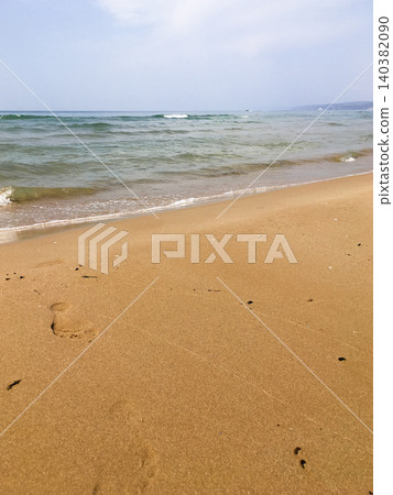 Waves softly lap against shore, creating serene ambiance on sandy beach. Footprints visible in golden sand, peaceful moment by water. Clear blue sea stretches out under light sky Waves softly lap against shore, creating serene ambiance on sandy beach. Footprints visible in golden sand, peaceful moment by water. Clear blue sea stretches out under light sky 140382090