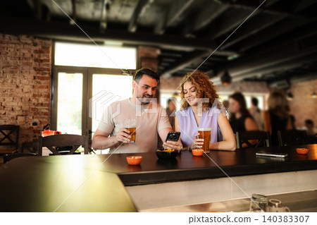 Couple enjoying beer and looking at smartphone at the bar counter Couple enjoying beer and looking at smartphone at the bar counter 140383307