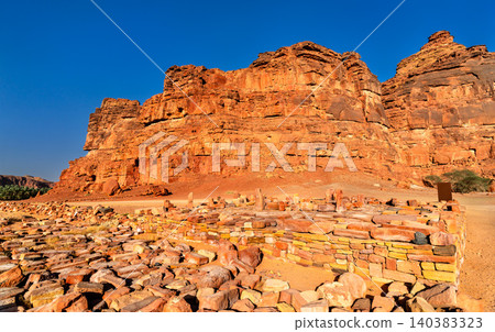 Ancient stone ruins at the Dadan archaeological site are set against massive red sandstone cliffs in AlUla, Saudi Arabia. 140383323