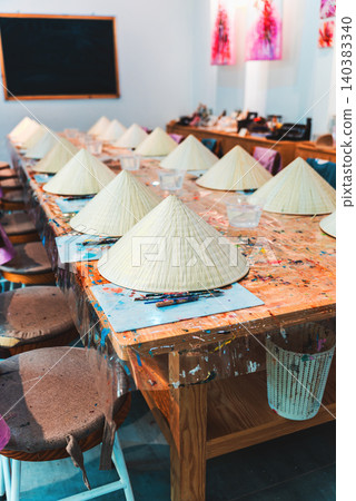 Non la hat painting workshop with brushes palettes and colorful paints on wooden table, Traditional Vietnam craft activity painting conical hats in creative art studio setting Non la hat painting workshop with brushes palettes and colorful paints on wooden table, Traditional Vietnam craft activity painting conical hats in creative art studio setting 140383340