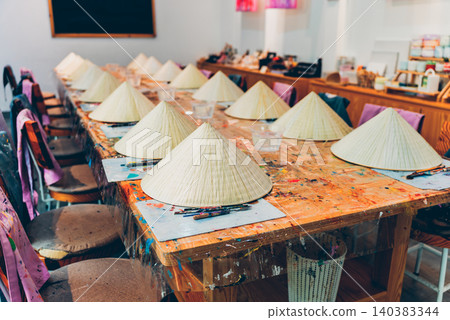 Non la hat painting workshop with brushes palettes and colorful paints on wooden table, Traditional Vietnam craft activity painting conical hats in creative art studio setting Non la hat painting workshop with brushes palettes and colorful paints on wooden table, Traditional Vietnam craft activity painting conical hats in creative art studio setting 140383344