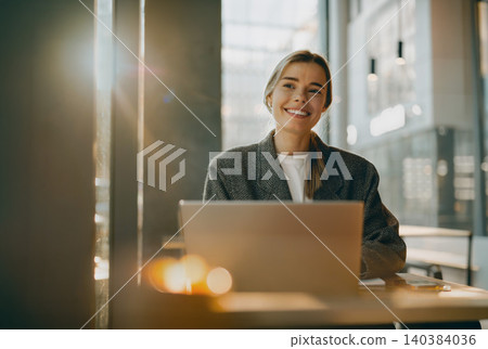 Happy young adult Caucasian woman smiling while using her laptop inside a bright modern cafe Happy young adult Caucasian woman smiling while using her laptop inside a bright modern cafe 140384036