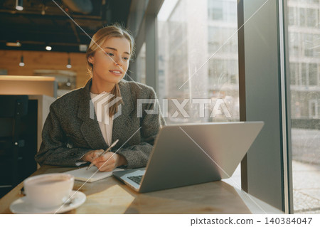 Caucasian young woman working on her laptop and writing in notepad inside a modern cafe Caucasian young woman working on her laptop and writing in notepad inside a modern cafe 140384047