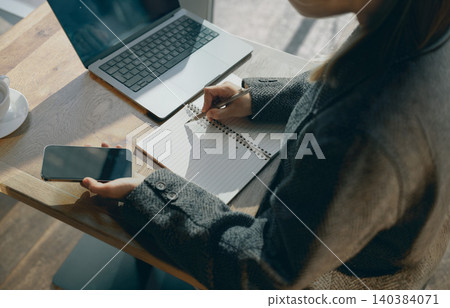 Asian young adult writing in notebook and holding smartphone near laptop in cafe Asian young adult writing in notebook and holding smartphone near laptop in cafe 140384071