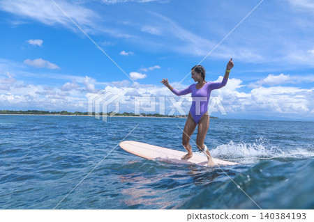 Lively Young Woman Guides Her Longboard Over Frothy Surf Under Blue Sky 140384193