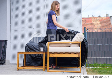 Woman removes black protective cover from wooden garden furniture on outdoor patio Woman removes black protective cover from wooden garden furniture on outdoor patio 140385023