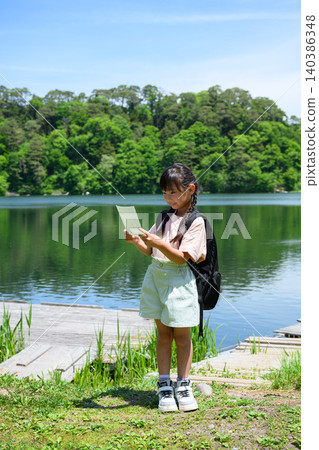 一名小學生在校外旅行時,在綠樹環繞的湖邊駐足,閱讀她的書籤。 一名小學生在校外旅行時,在綠樹環繞的湖邊駐足,閱讀她的書籤。 140386348