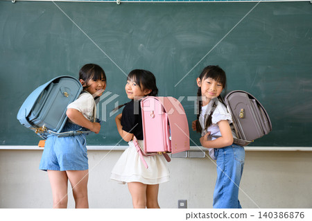 New girls, wearing colorful school bags, turn around with smiles on their faces in front of the classroom blackboard. New girls, wearing colorful school bags, turn around with smiles on their faces in front of the classroom blackboard. 140386876