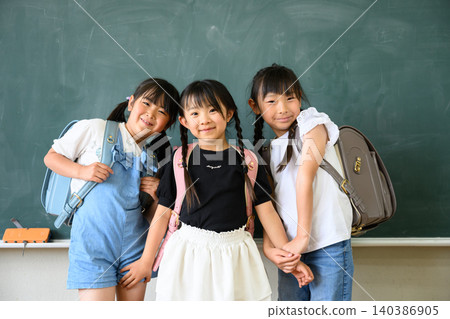 一群面帶笑容的小學生女孩手牽手,背著色彩鮮豔的書包,站在教室的黑板前。 一群面帶笑容的小學生女孩手牽手,背著色彩鮮豔的書包,站在教室的黑板前。 140386905