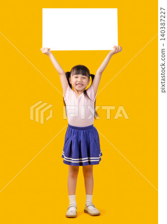Full length view of a cheerful little girl in a school uniform holding a blank white sign high above her head against a bright yellow background. 140387227