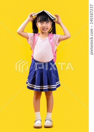 A cute Asian female student stands against a yellow background with a book on her head, happily and cheerfully welcoming the new semester. A cute Asian female student stands against a yellow background with a book on her head, happily and cheerfully welcoming the new semester. 140387237