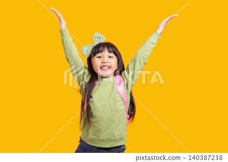 A happy Asian schoolgirl raises her hand with her backpack on; against a yellow background, the excited child exudes a sense of success and confidence. A happy Asian schoolgirl raises her hand with her backpack on; against a yellow background, the excited child exudes a sense of success and confidence. 140387238