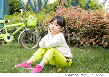 A lovely Asian girl sits on the grass outdoors, resting her chin on her hands, embodying a relaxed outdoor lifestyle. A lovely Asian girl sits on the grass outdoors, resting her chin on her hands, embodying a relaxed outdoor lifestyle. 140387252