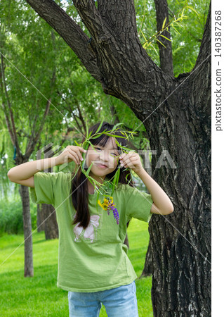 A playful young girl hiding behind a willow branch garland in front of a massive tree trunk. A playful young girl hiding behind a willow branch garland in front of a massive tree trunk. 140387268