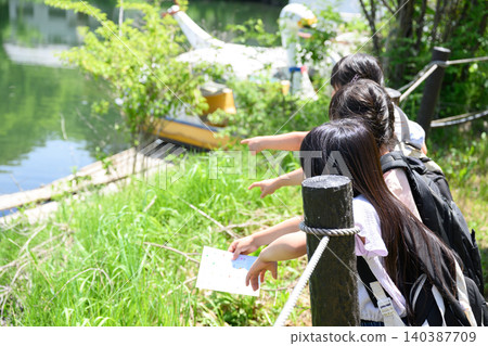 Elementary school girls with backpacks on stand by a lakeside surrounded by lush greenery, holding maps and pointing into the distance. Elementary school girls with backpacks on stand by a lakeside surrounded by lush greenery, holding maps and pointing into the distance. 140387709