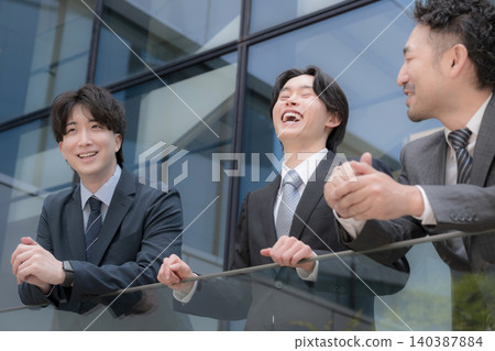 A team of company businessmen, smiling and looking towards the future with a building backdrop outdoors, discuss their growth, challenges, and project successes. 140387884