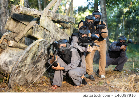 Paintball players of one team in camouflages and masks aiming with gun in shootout Paintball players of one team in camouflages and masks aiming with gun in shootout 140387916