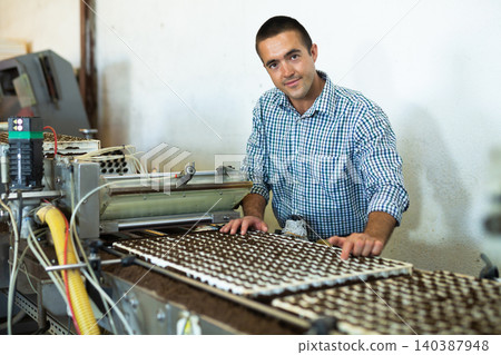 Farmer man working with automatic filling and seeding line for growing containerized seedlings of decorative plants in greenhouse 140387948