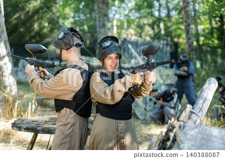 Boy and girl paintball players standing back-to-back and shooting Boy and girl paintball players standing back-to-back and shooting 140388087