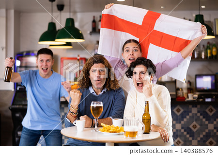 Company of young adult emotional fans supporting English team with state flag while resting in sports bar with beer Company of young adult emotional fans supporting English team with state flag while resting in sports bar with beer 140388576